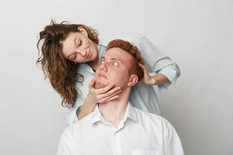 Young woman gently holds redhead man face with hands while looking at him. Couple in casual clothes on light background. Concept of love, care, relationship, and affection between partners.
