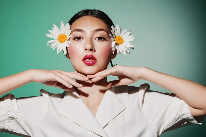 Young woman with bright makeup and two daisies in her hair poses with hands folded under her chin on a pastel green background. Creative concept in beauty fashion style.