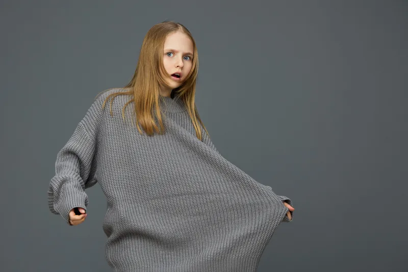 Surprised girl in oversized gray sweater against a neutral background, showcasing casual fashion and playful emotions, perfect for fashion or lifestyle themes