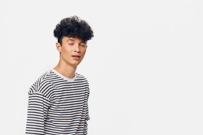 Young man with curly hair wearing striped shirt posing with closed eyes on white background, expressing calmness and relaxation in minimalist setting.