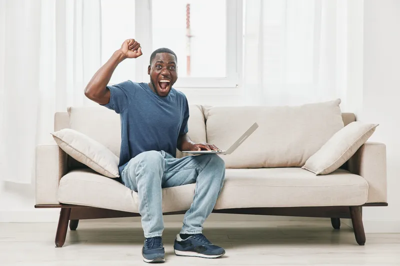 Excited man celebrating success while working on a laptop in a cozy living room setting with a light color palette