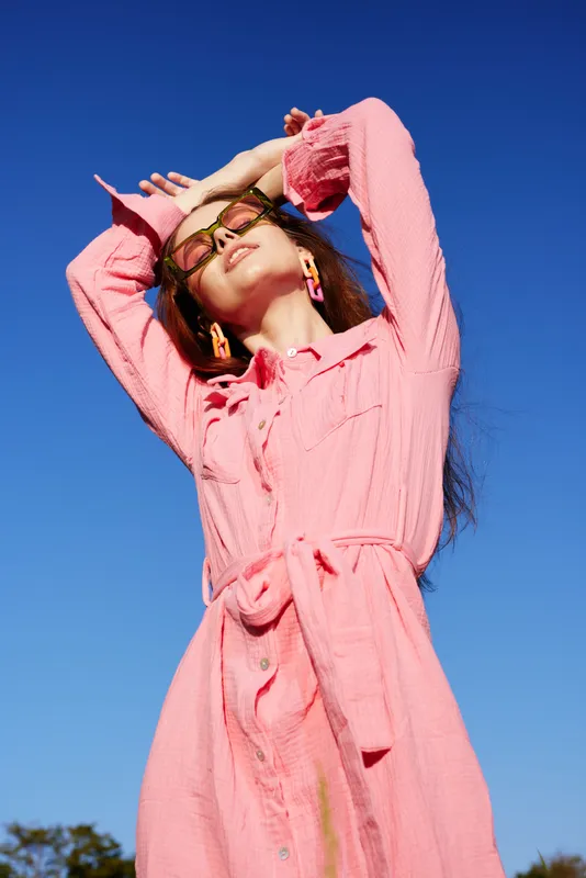 Young woman in a pink dress posing against a clear blue sky, showcasing vibrant summer fashion and joyful emotions, ideal for lifestyle and fashion photography