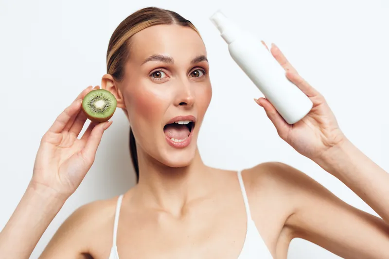 Young woman holds fresh kiwi slice and white cosmetic bottle shows natural skincare concept with healthy fruit and beauty product on white background with excited facial expression.
