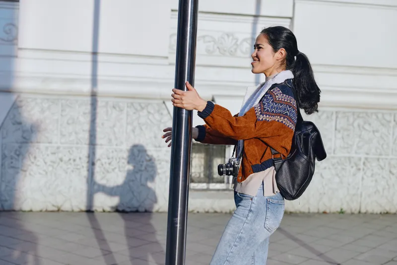 Woman clings to a street pole outdoors, wearing a brown orange sweater, scarf, jeans and backpack, with vivid urban light and a long shadow, conveying movement, casual style and city exploration