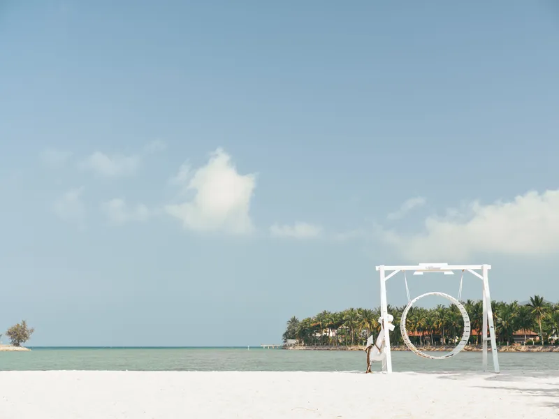 A serene beach scene featuring a white circular frame on pristine sand, calm turquoise sea, and a clear blue sky, with a lone figure nearby exploring the sculpture.