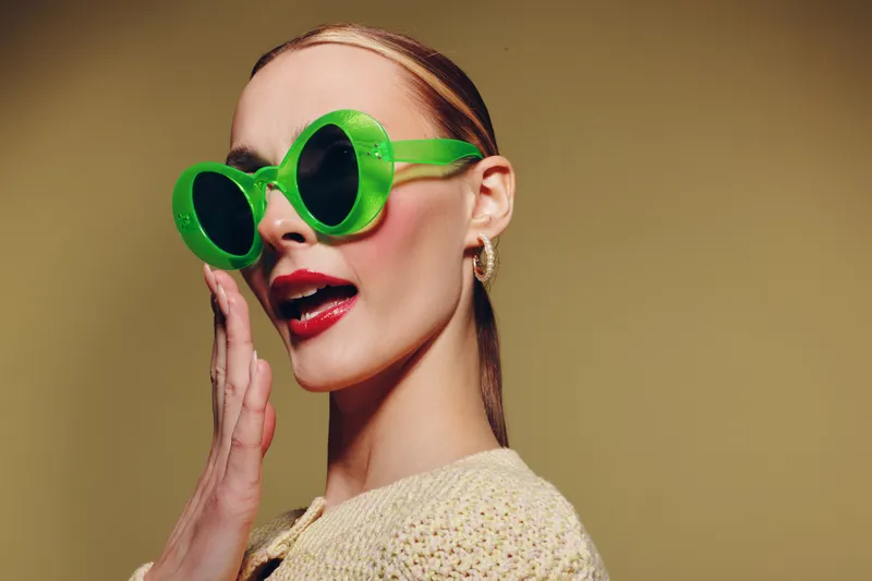 Stylish woman with green oversized sunglasses and red lipstick poses with hand near face on beige background. Concept of fashion, beauty, accessories, and retro style.