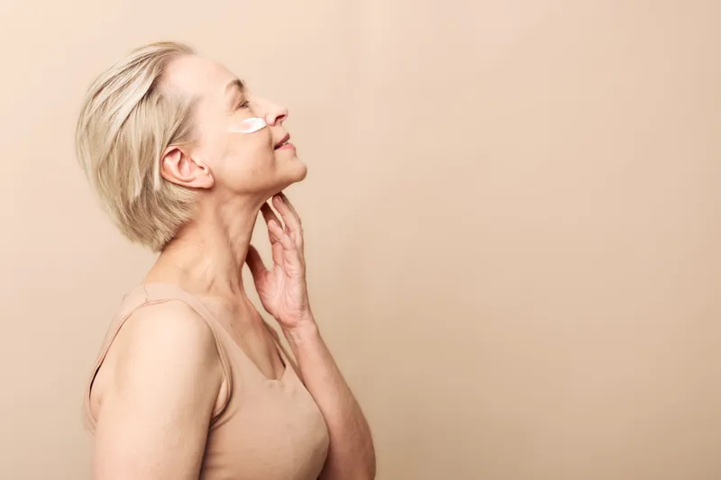 Mature woman with short blonde hair applies under eye patches in beige tank top. Side profile on neutral background. Concept of skincare, anti aging, and self care routine.