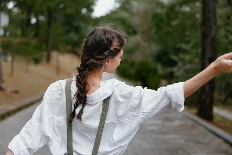 Young woman in casual outfit expressing freedom, with braided hair, walking in nature on a sunny day in a peaceful park setting, showcasing tranquility and happiness