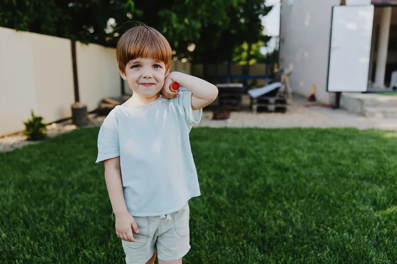 Joyful boy playing outdoors with a ball, smiling happily in a sunny backyard filled with greenery and playful ambiance. Perfect for family-related themes.