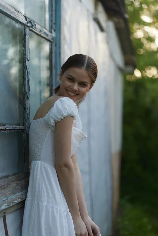 Young woman in a white dress smiling near an old wooden house, surrounded by greenery, evoking a feeling of happiness and nostalgia during summer