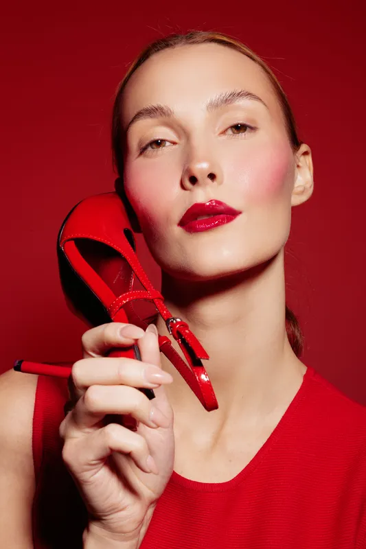 Close-up portrait of a woman with red lipstick holding a red high heel shoe near her face against a red background. Concept of fashion, beauty, style, and elegance.