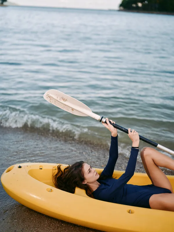 Relaxing woman lying in a yellow kayak on the beach, holding a paddle with calm sea waves in the background on a sunny day.