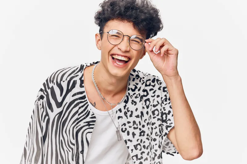 laughing man wearing glasses with curly hair in stylish black and white animal print shirt, joyful expression and casual white background, youthful happiness