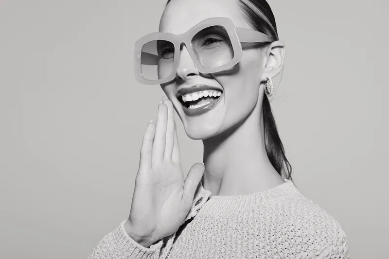 Smiling young Caucasian woman with light skin wearing large sunglasses and a knitted sweater, posing in studio. Fashion and style concept.