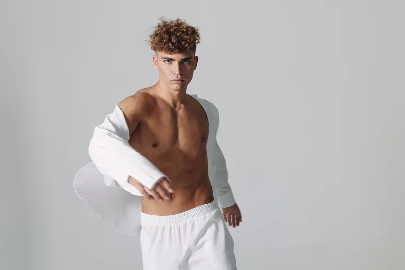 Young man with curly hair posing shirtless in white pants and open white shirt against plain background. Fashion model with confident expression and athletic body in studio lighting.
