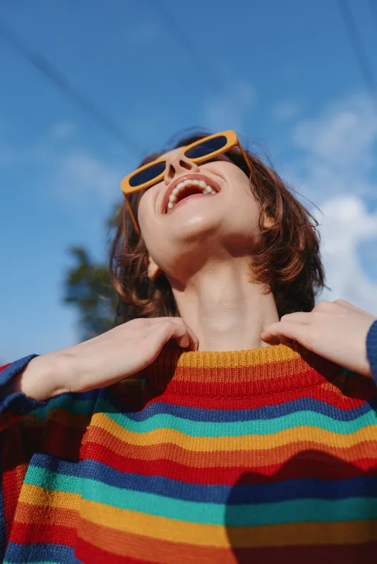 Woman smiling in colorful striped sweater and yellow sunglasses outdoors, young adult model laughing and looking up, casual outfit, joyful mood, bright blue sky, lifestyle travel vibe
