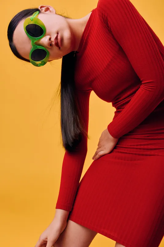 Young woman wearing a red dress and green sunglasses posing against a yellow background. Stylish female model with long dark hair and trendy fashion accessories in studio light.