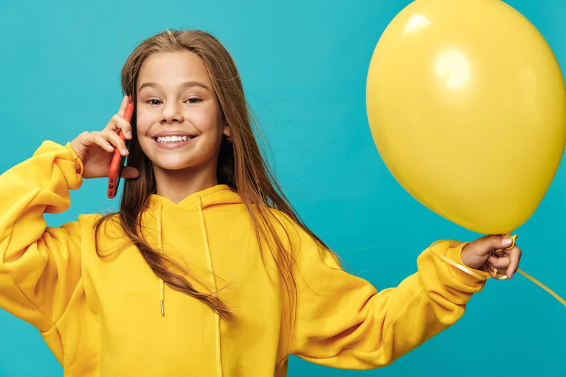 Girl in yellow hoodie smiling and talking on phone while holding yellow balloon with blue background. Child with long hair enjoying communication and celebration indoors.