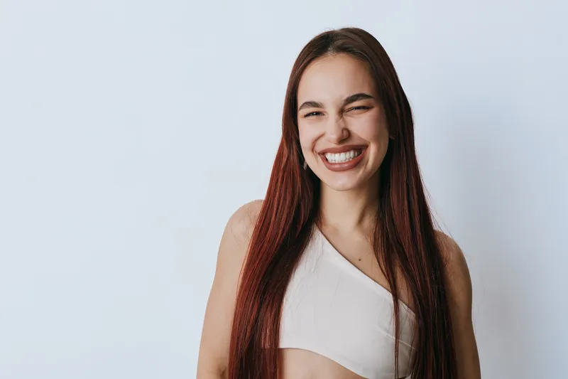 Young woman with long red hair smiling with eyes closed in casual white top against light background. Portrait of happy female expressing joy and positive emotion indoors.