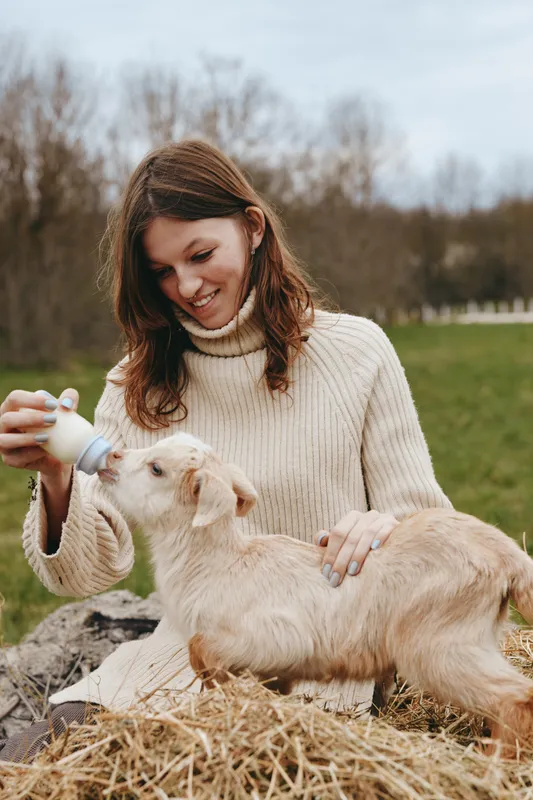 Smiling woman bottles feeds a goat kid outdoors in a field, portraying a lifestyle moment of care, bonding, and gentle interaction with animals.