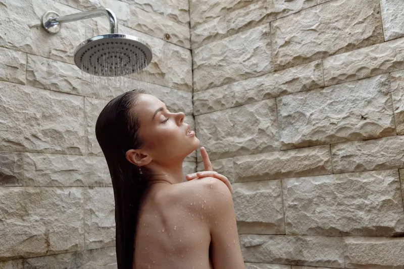 woman showering indoors with wet hair under rain showerhead on stone textured wall, relaxing and refreshing moment in bathroom setting