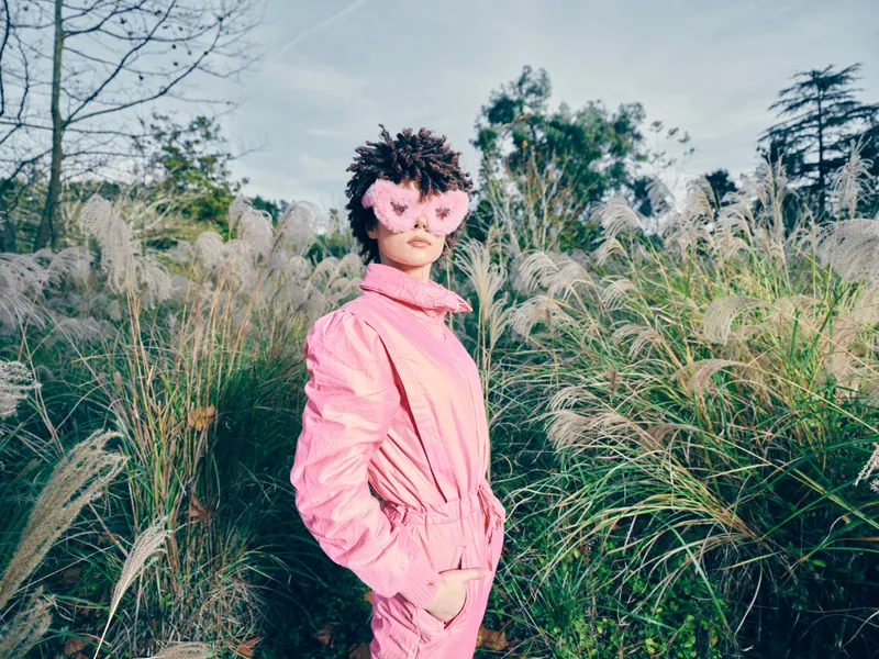Pink outfit and masked person pose in a field of tall grasses, bold color contrasts with greenery, outdoor fashion scene showcasing mystery, drama and artistic vibe in nature