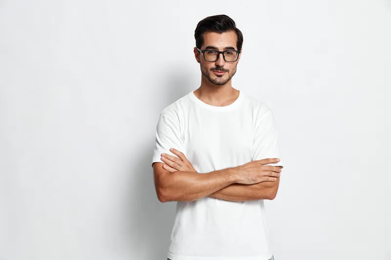 Confident young man with glasses standing with arms crossed, wearing a white t-shirt, looking at the camera against a solid light background, studio portrait style.