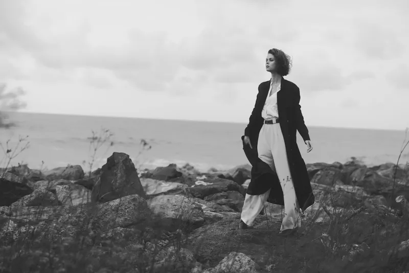 Woman in a long coat walks along a rocky coast in monochrome, the sea and horizon framed by rugged scenery and moody daylight