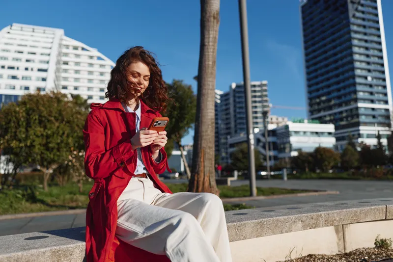 Young woman with curly hair sits on a concrete ledge in a red coat, reading her phone in an urban park as bright buildings rise behind palm trees.