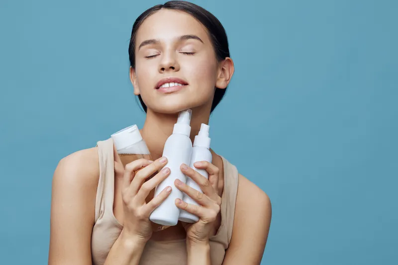 Young woman with closed eyes joyfully holding skincare products on a blue background, showcasing a feeling of serenity and self care with a focus on beauty and health