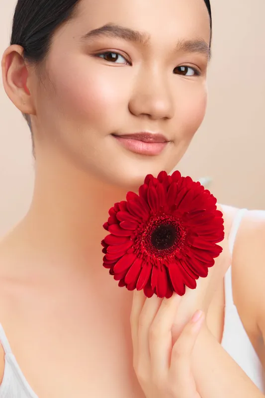 Asian woman holds a bright red gerbera flower in a close-up portrait, soft lighting and gentle expression create a calm, elegant beauty shot suitable for lifestyle and wellness themes.