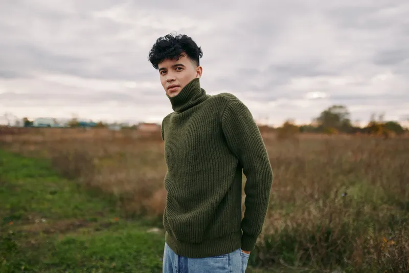 Portrait of a young man in a knit turtleneck standing in an open field, natural pose and authentic presence conveying emotional authenticity and quiet veracity in a simple rural scene.