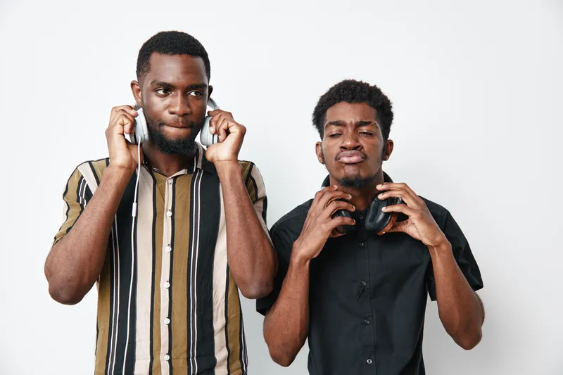 Two young black men with dark skin holding headphones near ears on white background. Casual clothing, facial expressions showing focus and enjoyment in studio.