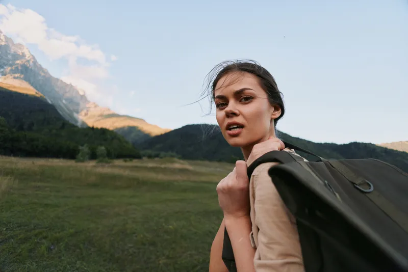 Young woman with backpack traverses a grassy valley beneath rugged mountains, turning her head with a determined, alert gaze, wearing a light jacket and sturdy shoes.