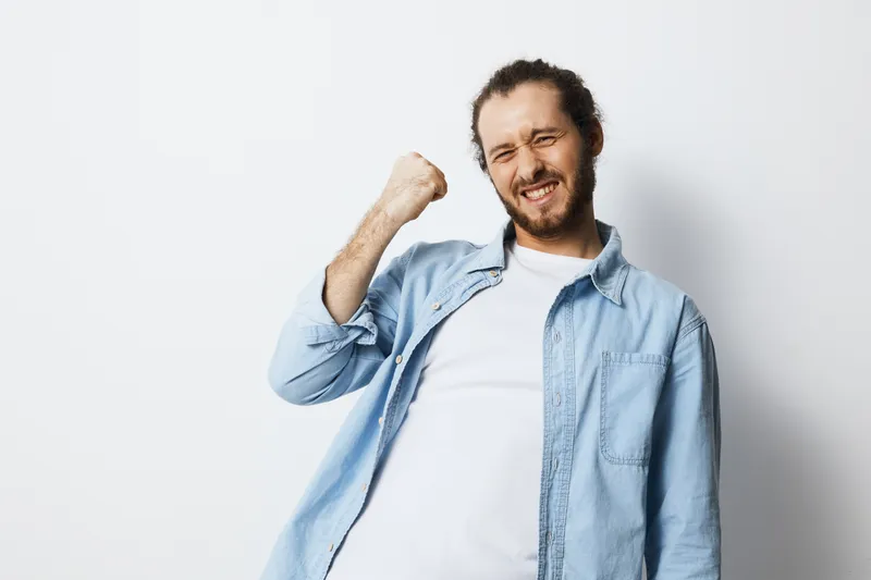 A joyful young man celebrating success with a raised fist, wearing a light blue shirt against a minimalist white background Emphasis on positivity and achievement