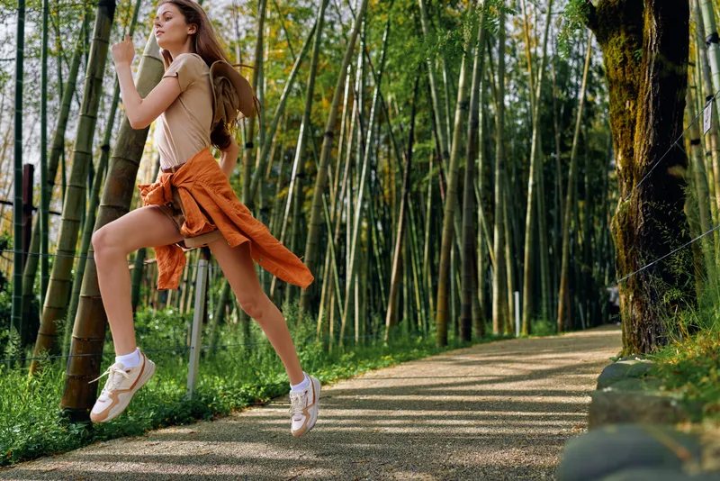 Woman jump on forest path with backpack and orange jacket tied at waist, joyful motion and sneakers. Outdoor bamboo grove on walking trail, travel, fitness and adventure energy.