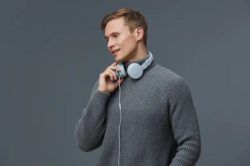 Stylish young man wearing gray knitted sweater smiling listen to music with over-ear headphones on gray background. Casual fashion, lifestyle, leisure, happiness, modern attire, indoor setting.