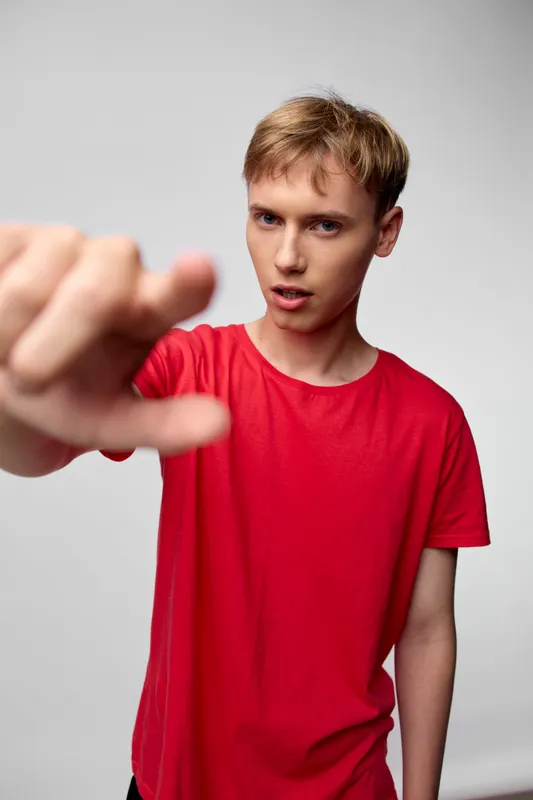 Close up of a young man in a red shirt reaching toward the camera, with a determined expression and focused gesture, studio lighting, conveying action and engagement in a dynamic pose.