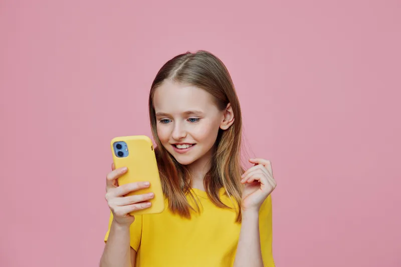 Young girl in a yellow t shirt, smiling while using a smartphone against a pink background, expressing joy and curiosity, ideal for social media concepts