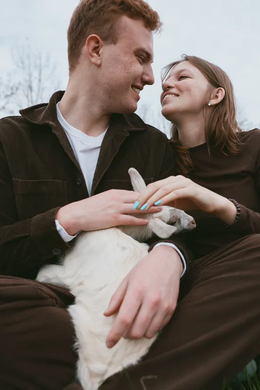 Joyful couple with a goat kid outdoors, sharing a warm affectionate moment in a lifestyle portrait. Natural light highlights connection happiness and wellbeing in a calm rustic setting.