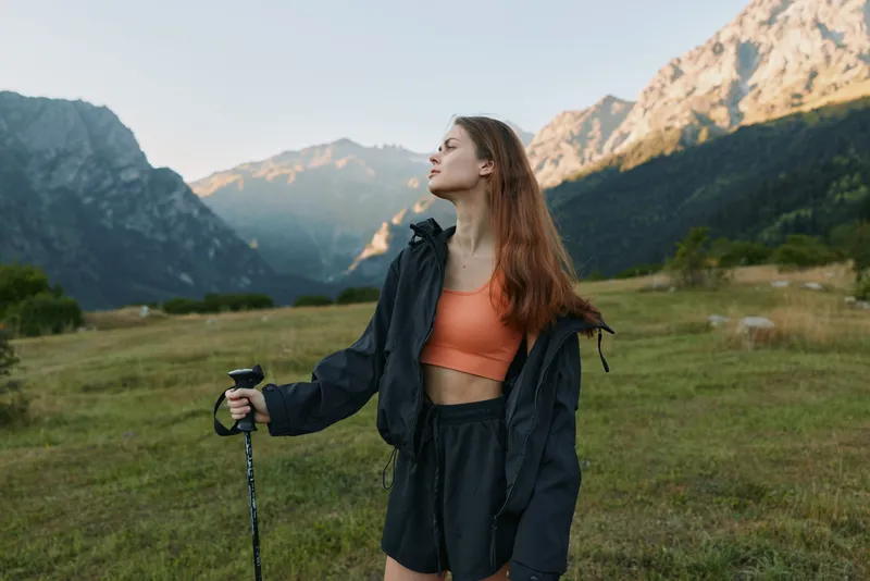 A confident young woman stands in a grassy valley with a trekking pole, wearing a windbreaker and shorts, framed by towering mountains under clear skies, and a light breeze.