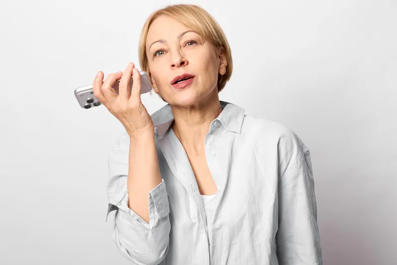 Stressed mature woman in white shirt holds phone handset near ear, worried expression, studio portrait on plain white background, communication call concept