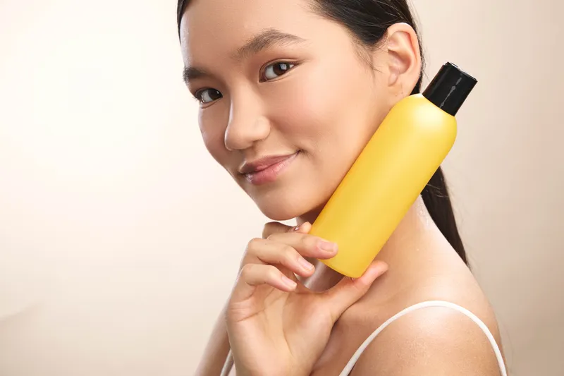 Asian woman holds a yellow bottle close to her face in a soft studio setting, smiling with confidence, conveying a bright skincare moment and personal care appeal.