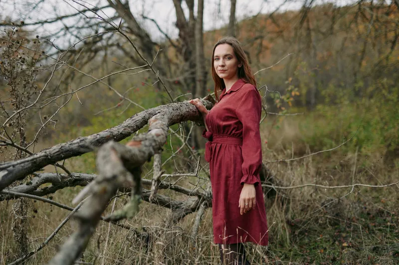 A candid portrait of a woman in a red dress standing beside a fallen tree in a wild meadow, conveying authentic presence, natural poise and quiet confidence in soft autumn light.