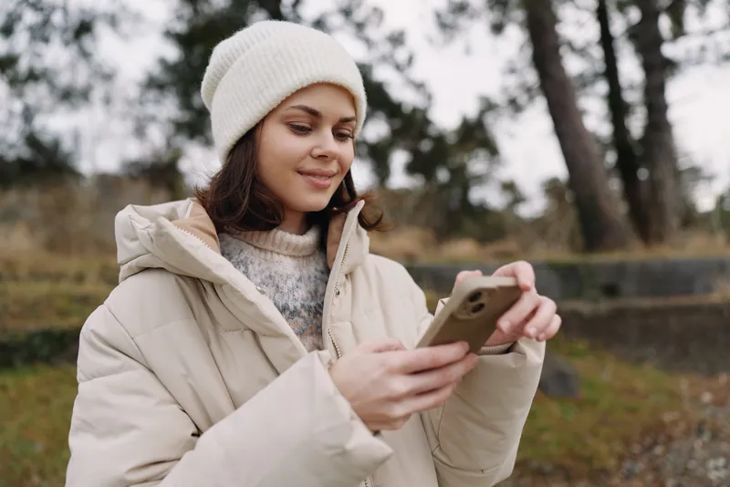 Winter scene with a woman checking a smartphone outdoors, wearing a cozy jacket and knit beanie, in a park setting on a chilly day, capturing casual lifestyle and digital connection.