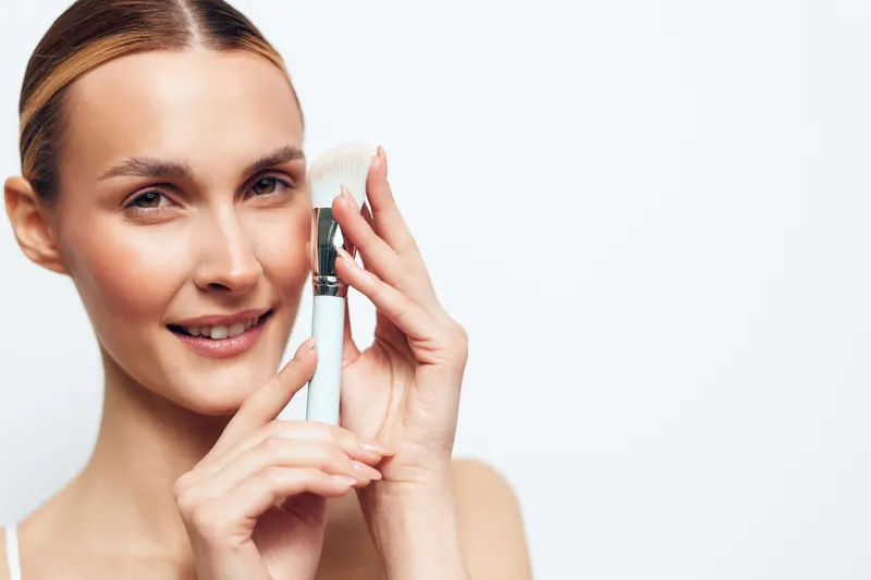A smiling young woman with clear skin holds a white makeup brush near her face. The bright studio background highlights skincare and beauty concepts with natural light and soft focus.