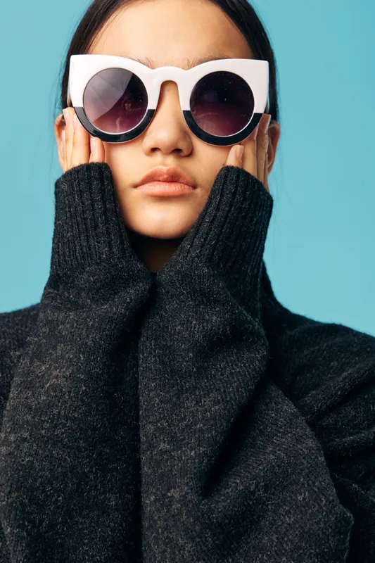 Woman with large sunglasses and black sweater posing against blue background, fashion portrait with cozy outfit and stylish eyewear for casual modern look.