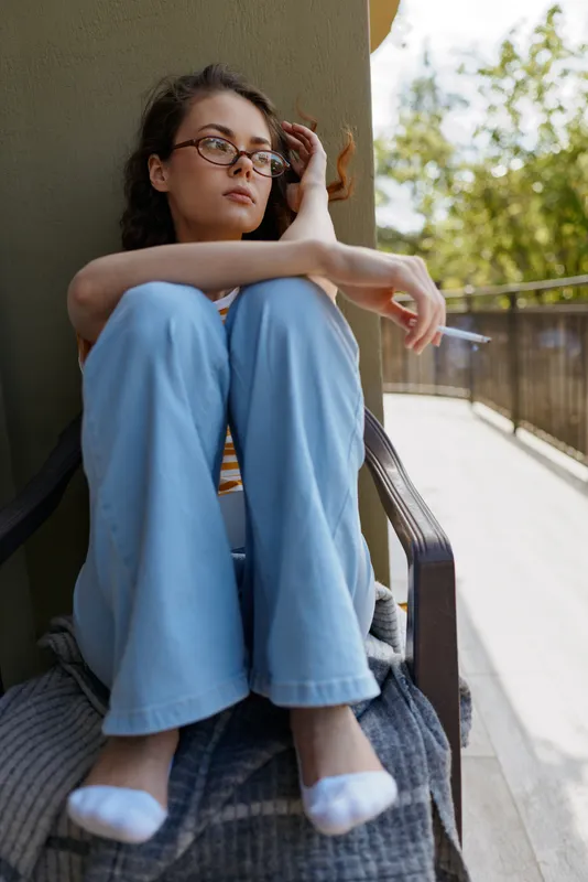 Young woman with glasses sits on a balcony in a casual home setting, leaning against a railing and gazing thoughtfully into the distance as warm daylight brightens her relaxed pose.