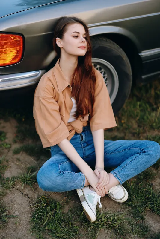 Young woman sitting outdoors by a vintage car, wearing casual fashion with a relaxed expression, capturing a serene moment in a natural setting.