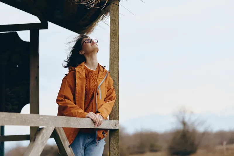 Woman lifestyle outdoors portrait, leaning on wooden fence, wearing orange jacket and eyeglasses, curly hair tied back, calm thoughtful expression, soft spring light, open sky background